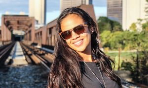 A woman with long dark hair and sunglasses smiles outdoors on a sunny day.