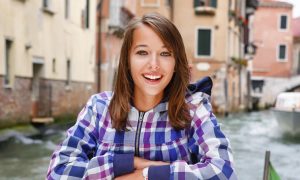 A smiling woman in a purple plaid shirt sitting outdoors in a city.