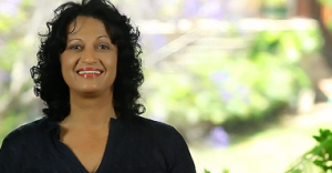 Smiling woman with curly hair in a dark top against a blurred outdoor background.