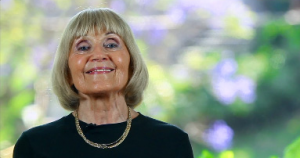 Smiling elderly woman with blonde hair and a gold chain necklace.