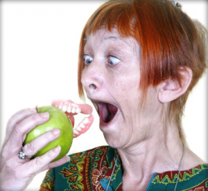 A surprised red-haired person holding a green apple with fake vampire teeth.