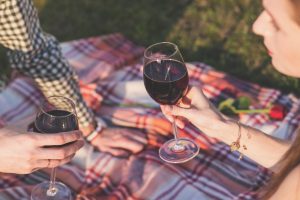 Two people clinking wine glasses on a picnic blanket outdoors.