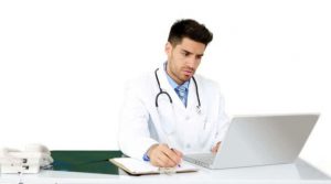 Young male doctor writing notes with a laptop nearby.