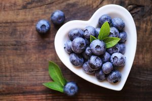 Fresh blueberries in a heart-shaped bowl on wooden surface.