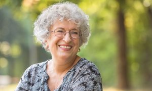 Smiling elderly woman with curly gray hair and glasses outdoors.